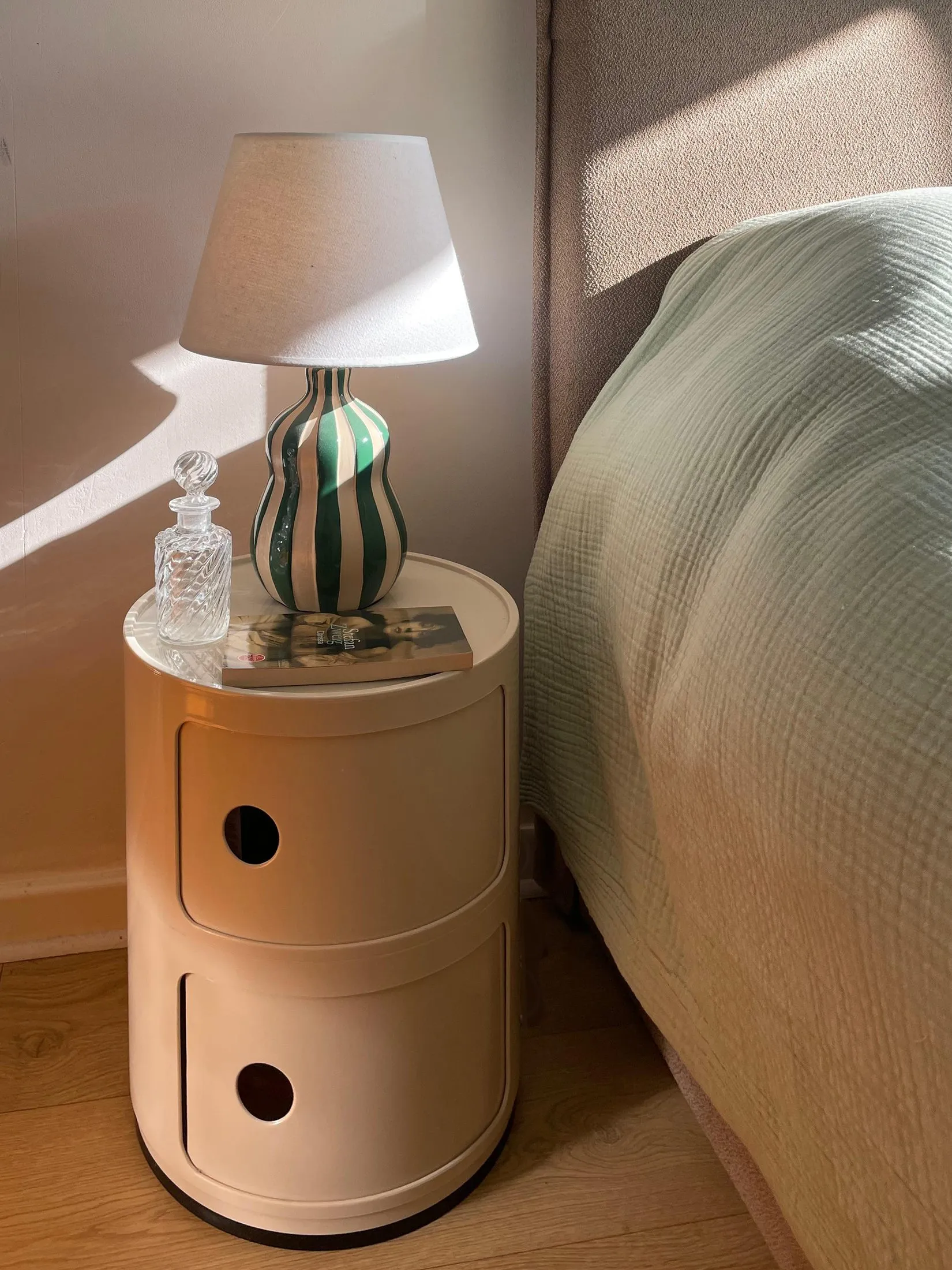 Table de chevet cylindrique beige et lampe à pied vert, contre un lit aux draps vert d'eau
