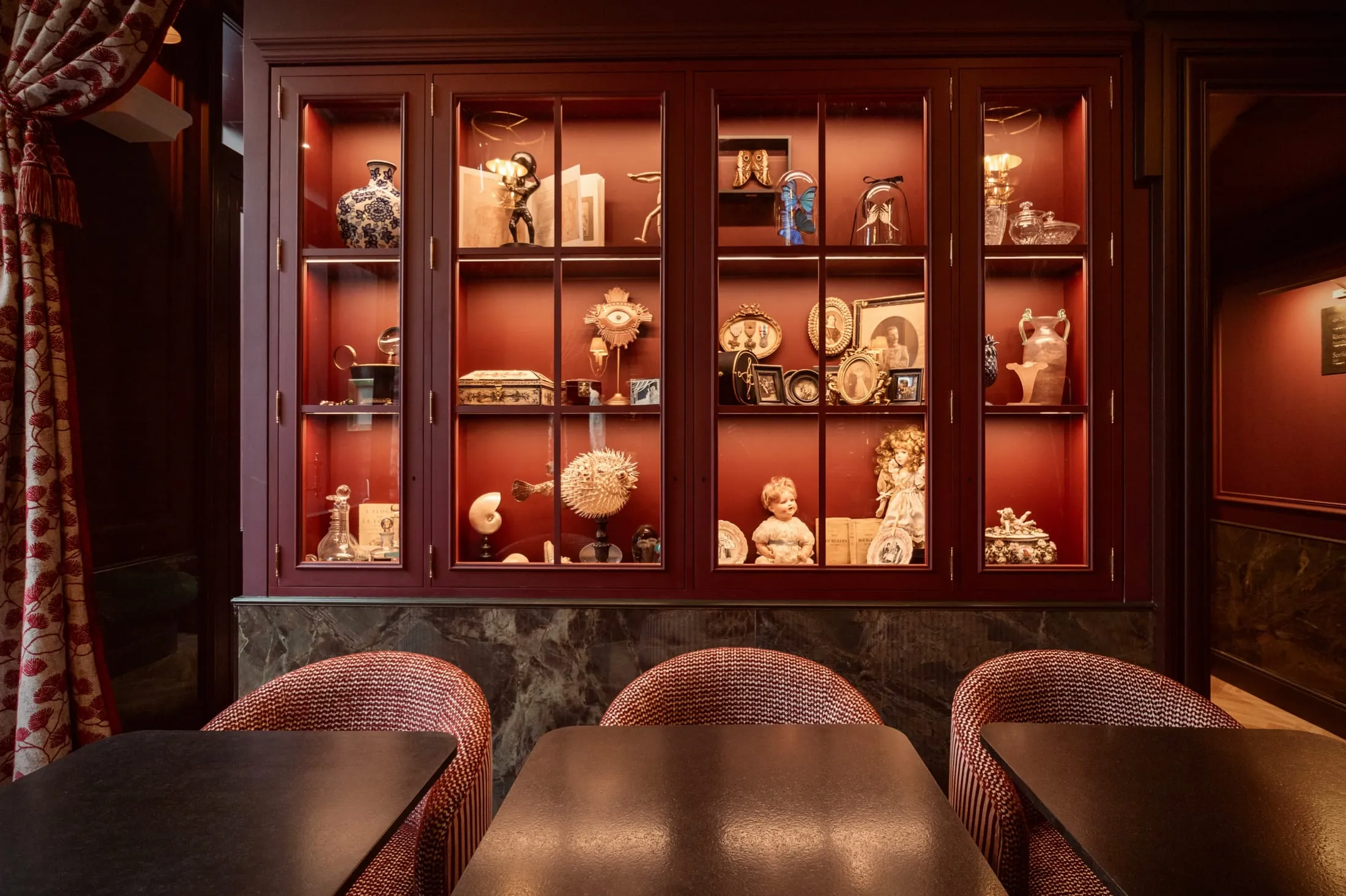 Vitrine de curiosités éclairée au fond du salon, fauteuils cannés au premier plan